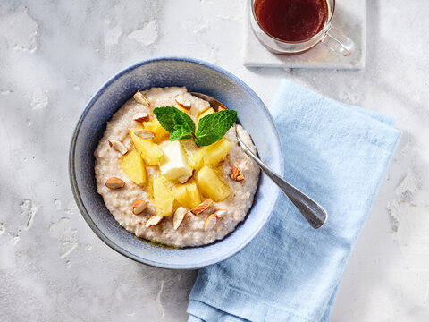 Warm oatmeal is topped with juicy pineapple chunks and sliced almonds, garnished with mint leaves. A cozy cup of herbal tea is beside it on a light colored countertop