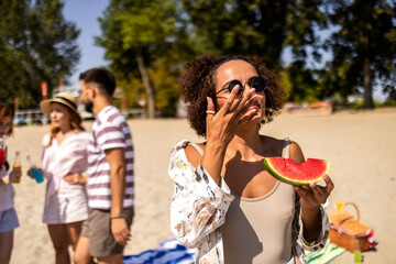 Obraz premium African American young woman eats watermelon at the beach while her friends, behind her, drink cocktails and talk.