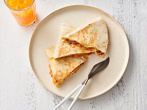 Three cheesy quesadillas cut into triangles sit on a neutral plate next to a glass of orange juice. The bright kitchen setting adds warmth to the meal