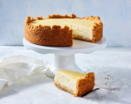 A creamy cheesecake sits enticingly on a white cake stand, showcasing its smooth texture and golden crust. A slice is cut out, inviting dessert enthusiasts to indulge