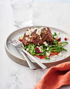 Juicy steak rests atop a bed of fresh arugula, garnished with toasted pine nuts and dried cranberries, creating a vibrant and appetizing dish perfectly arranged on a sleek plate