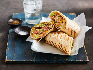 Three tasty wraps filled with greens, meats, and spices are presented on a rustic plate with a glass of water nearby. Small bowls with salt and seasoning add flavor