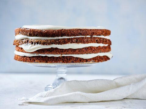 A delightful layered cake sits on a glass stand, featuring three layers of rich brown cake separated by smooth white frosting, ideal for special occasions or gatherings