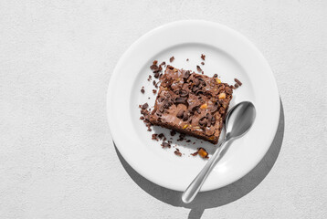 Chocolate brownie with nuts on a plate with spoon on a light background with morning shadow.