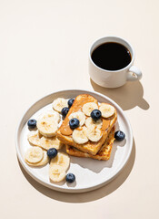 Peanut butter toast with banana and blueberry on a plate on a beige background with a cup of espresso coffee