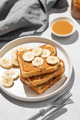 Peanut butter toast with banana on a plate on a light background with napkin and shadow.