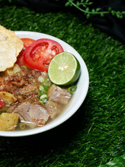 Close up clear beef soup with sliced beef, potatoes, tomatoes, lime, sprinkled with spring onions, and fried onions. Served in a white bowl with a green synthetic grass background.