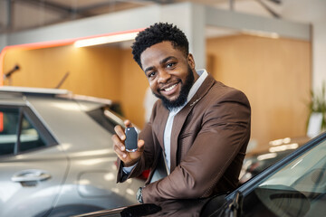 Portrait of smiling african american car seller leaning on brand new luxurious car and showing car...