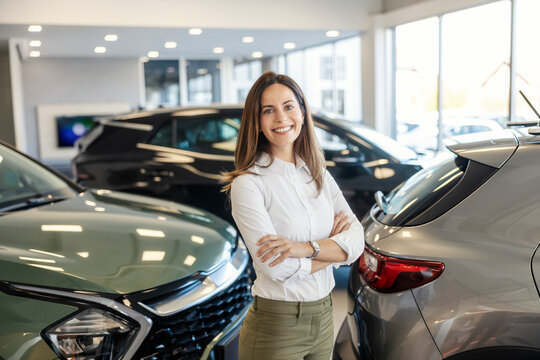 Portrait of happy confident car saleswoman standing at car showroom with arms crossed and smiling.