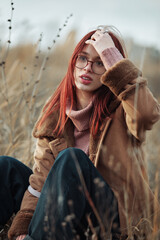 Teenage girl with red hair and glasses sitting in tall grass, touching her hair with a thoughtful...