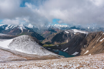 Scenic view from above to most beautiful turquoise alpine lake among hills in freshly fallen snow and large snowy mountains in low clouds. High mountain range with snow-white peaks in low cloudy sky.