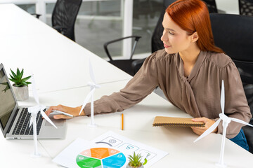 Young female engineer sitting at the table and looking at the wind turbine model