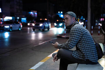 An Asian Man Waiting for a Bus After Working Late, Using His Smartphone at a Busy City Bus Stop, Capturing the Struggles of Urban Life, Overwork, and the Nightly Commute in a Chaotic Capital