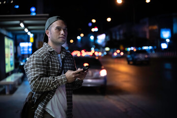 An Asian Man Waiting for a Bus After Working Late, Using His Smartphone at a Busy City Bus Stop, Capturing the Struggles of Urban Life, Overwork, and the Nightly Commute in a Chaotic Capital