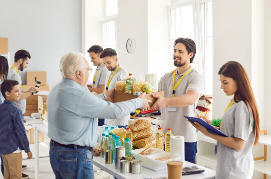 Happy volunteer at the aid center gives an old man a box of food, close-up. Group of volunteers working on a humanitarian project. Concept canned food, humanitarian aid, non-profit organization - Powered by Adobe