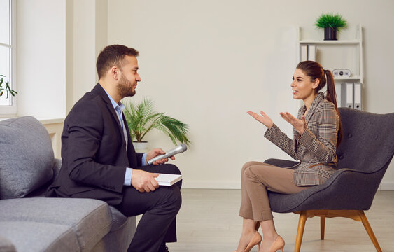 Portrait of male young journalist in suit with microphone in hand interviewing attractive smiling woman sitting on chair indoors. Man talking with his female guest during interview in office.