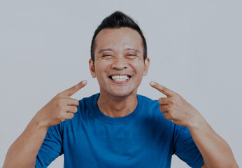Asian man showing his big smile and pointing at his tooth, isolated over white background.
