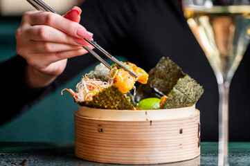 A close-up of a hand using chopsticks to pick up colorful sushi rolls and garnishes from a bamboo...
