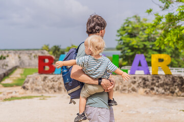 Father and son tourists posing in front of the colorful BACALAR sign with vibrant letters, trees, and Bacalar Lake in the background. Mexican travel and adventure concept