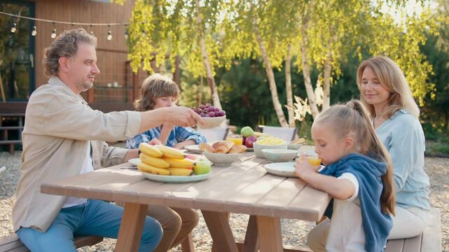 Camera moving around table where Caucasian family enjoying healthy breakfast. Little girl drinking fresh juice while father passing bowl of grapes to beloved wife. Green bushes in background.