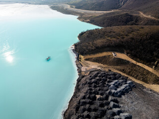 A turquoise tailings pond near a cement factory, surrounded by rugged hills. The vivid water...