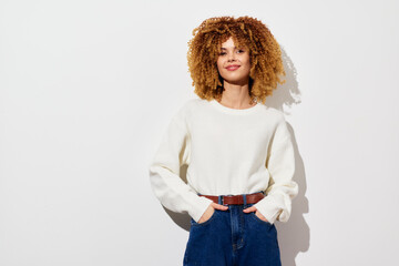 Smiling young woman with curly hair wearing a cozy white sweater and high waisted jeans against a minimalistic white background, exuding confidence and warmth