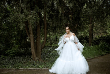 a beautiful bride in a white dress and a long veil stands against the background of dark greenery