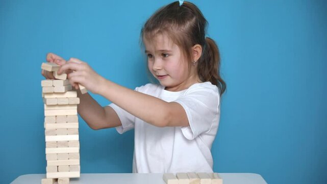 Slow motion footage little girl playing a jenga board game on blue background. Happy child takes wooden blocks from tumble tower and it's falling down. Children entertainment and creativity concept. 