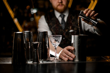 Selective focus on male bartender's hand pours liquid from cocktail jigger into metal glass
