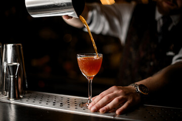 Hands of a bartender pouring a brown cocktail from a mixing glass into a wine glass