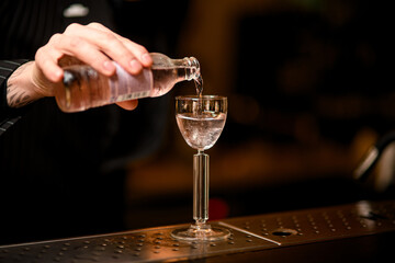 Focus on stemmed glass, into which a male bartender hand pours vodka