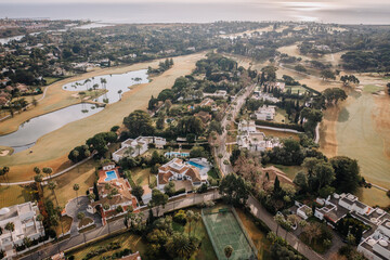 Aerial view of luxury villas, tennis courts, swimming pools, golf courses, and a scenic coastal area with water features and palm trees. Drone shot.