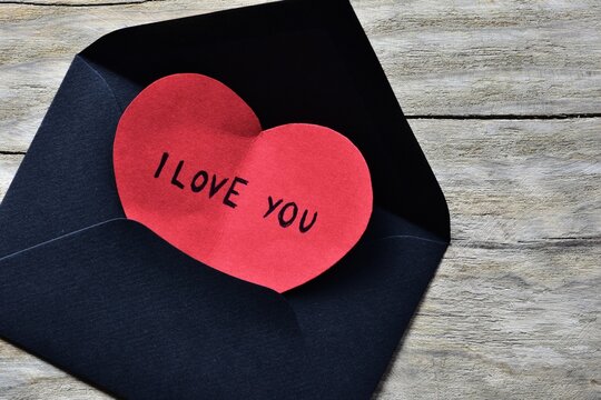 Message text I love you on red heart shape paper card for valentines day in small black envelope on wooden board textured background. Valentines day card with words I love you.