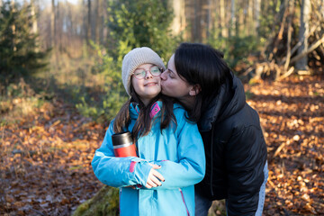 A cute little German girl with glasses and a thermos in her hands stands in the middle of an autumn forest being kissed by her mother. Active family pastime concept