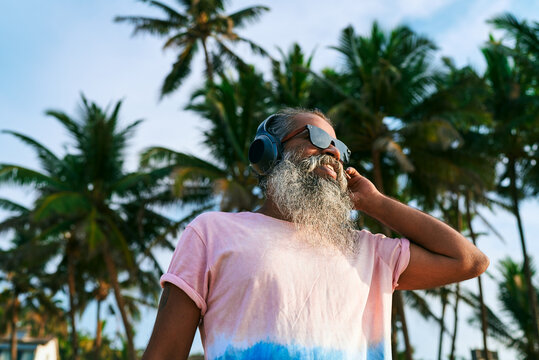 Older man with beard wears headphones, sunglasses near palm trees. Enjoys music experience. Maybe tries active noise cancellation. Tropical setting. Calm and relaxed mood.