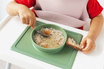Close-up of a child s hands holding a spoon and a slice of bread while eating chicken noodle soup from a green bowl, illustrating a wholesome and comforting meal