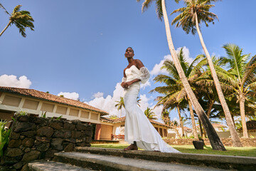 Ambiguous gender person in white wedding dress at tropical villa. Stylish bride on steps among palm...