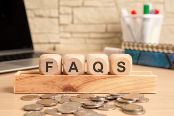 wooden blocks spelling FAQS, placed on a desk with coins, books, and stationery in a office setting