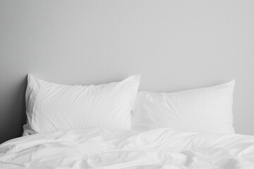 A neatly made bed with two fluffy white pillows against a light gray wall. The bedding is white and slightly rumpled, creating a cozy atmosphere.