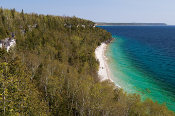 A lookout view of the deep blue and turquoise waters of a rocky shoreline, cliffs, and a cape point, surrounded by lush forests. 