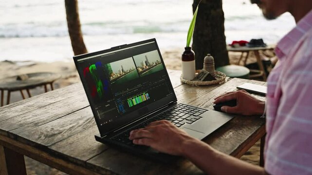 Blogger at beach cafe edits video on laptop, color grading sunny ocean footage. Freelancer working remotely, enhancing travel film during beautiful sunset. Digital nomad lifestyle at seaside. Slowmo