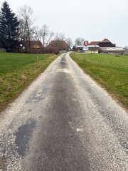 A dirt road stretching through a green countryside landscape under a cloudy sky. Power lines are visible on the left, adding contrast to the natural surroundings. The scene captures the calm and