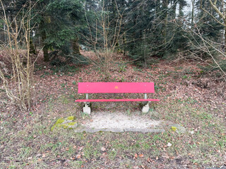 A vibrant red bench placed in the middle of a forest. Surrounded by tall trees and natural...