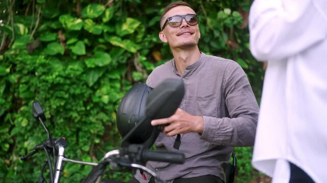 Nice happy couple puts on motorcycle helmets gets on motorbike ready to start off. People dress protection for bike, sit on scooter to go on trip in jungle. Man woman comply safety rules.