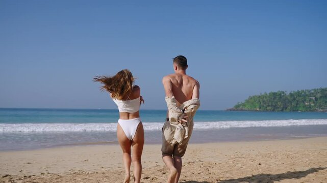 Young man and woman taking off clothes and throwing it up, running to the ocean. Fit couple having fun on summer vacation on the tropical sandy beach. Lovers enjoy romantic journey.