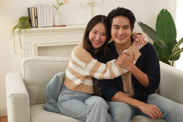 Obraz premium Asian couple, wife hugging husband, sitting on sofa together happy in living room at home, creating a warm and loving family in a new home.