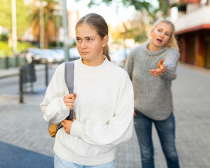Frowning teenage girl walking along city street on spring day, listening to reprimanding from her mother ..
