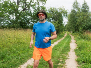 Fototapeta premium A man in a blue shirt and orange shorts walks down a dirt road. The man is wearing a hat and he is enjoying the outdoors. Male in his 40s with grey beard. Sport and fitness outdoor.