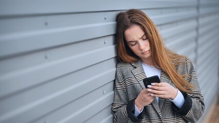 Sad young businesswoman leaning against a metal wall, reading distressing news on her smartphone,...