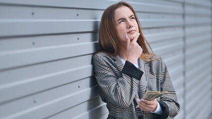 Serious businesswoman contemplating financial strategy, clutching dollar bills near weathered metal...